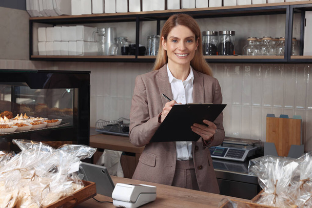 Happy business owner with clipboard and pen at cashier desk in bakery shop - Foto, Imagen
