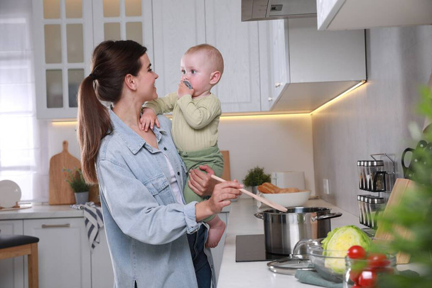 Happy young woman holding her cute little baby while cooking in kitchen - Fotoğraf, Görsel