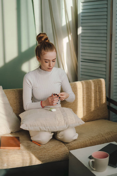 Mujer joven positiva sentada en un sofá y leyendo un libro interesante mientras descansa en la acogedora sala de estar en casa - Foto, Imagen
