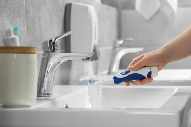 Woman washing electric toothbrush under flowing water from faucet in bathroom, closeup - Valokuva, kuva