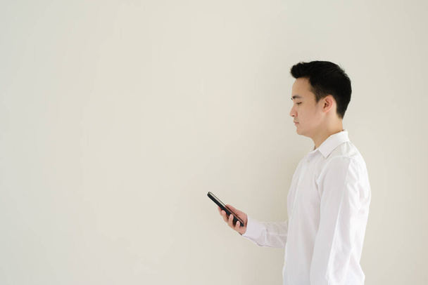 Beyaz tişörtlü genç bir Asyalı sağ tarafına bakarken akıllı telefona bakıyor. İzole edilmiş beyaz arkaplan. - Fotoğraf, Görsel