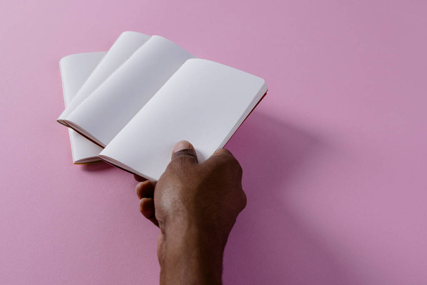 Hand of biracial man holding notebook over notebooks with copy space on pink background. Literature, reading, leisure time and books. - Φωτογραφία, εικόνα
