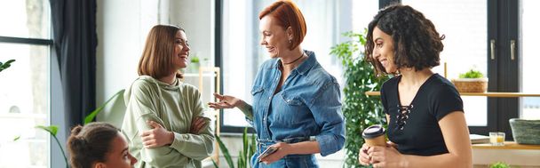 redhead woman with coffee to go pointing with hand while talking to carefree friend near multiethnic members of interest club, happy gathering and leisure of diverse female team, banner - Photo, Image