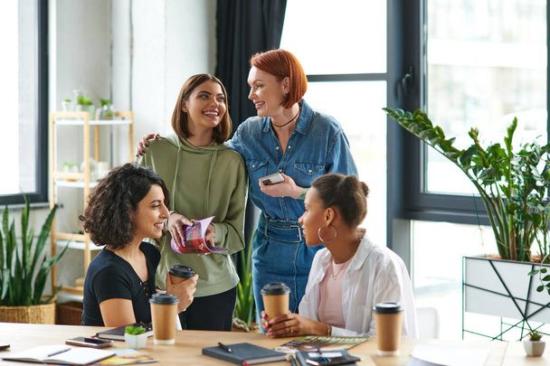 happy redhead woman with smartphone embracing young female friend near multiethnic women drinking coffee to go during conversation n interest club, solidarity and understanding concept - Photo, Image