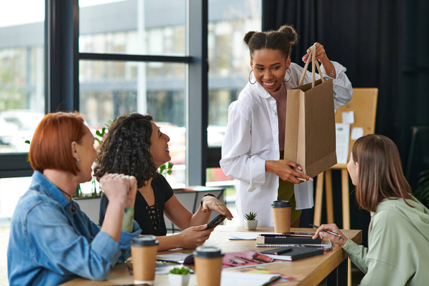 young, smiling african american woman showing shopping bag to diverse group of multiethnic female friends sitting near paper cups and notebooks in interest club, sharing joy and positive emotions  - Photo, Image