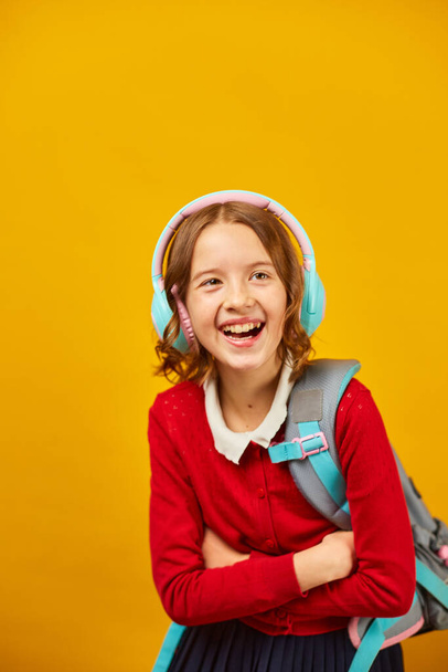 ハッピー女子高生バックパック,ヘッドフォンを持って楽しい休日,学校は夏のために黄色のスタジオの背景に,十代の女の子で学校の制服. - 写真・画像