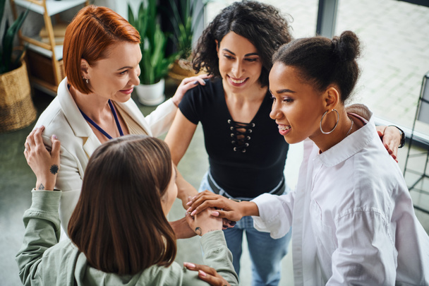 high angle view of joyous multiethnic girlfriends and smiling redhead psychologist joining hands as sign of unity during motivation session, moral support and mental wellness concept - Photo, Image