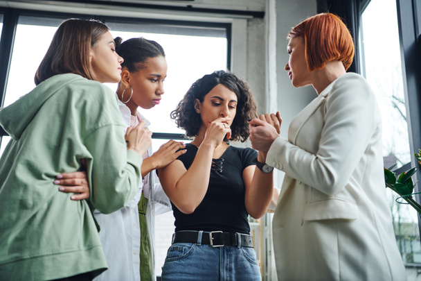 psychologist holding hand of frustrated multiracial woman and calming her together with multiethnic friends during motivation session in consulting room, problem-solving and mutual support concept - Photo, Image