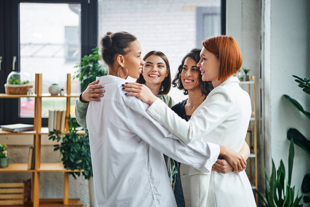 переполненные радостью многоэтнические женщины и рыжая мотивация тренер обнимать и смотреть друг на друга, стоя вместе в консультационной комнате, женское единство и концепция поддержки - Фото, изображение