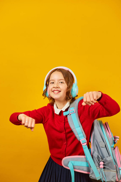 Happy schoolgirl with backpack, headphone having fun holiday, School's out for summer on yellow studio background, Teenager girl in school uniform. - Φωτογραφία, εικόνα