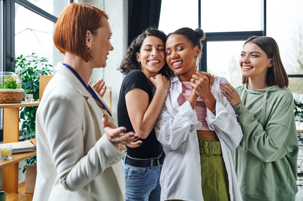 cheerful multiethnic women embracing african american girlfriend near happy motivation coach gesturing in consulting room, female unity and support concept - Photo, Image