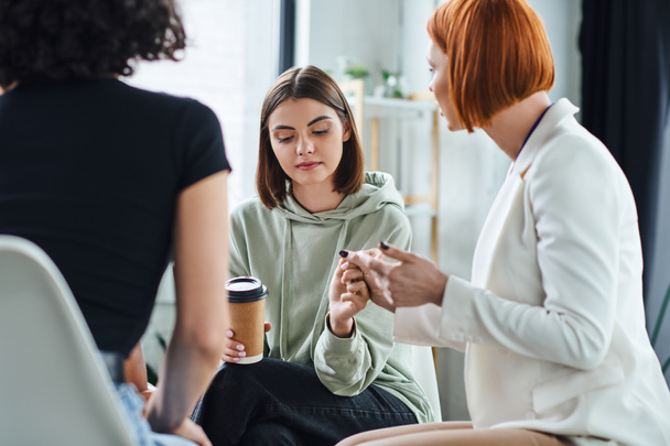 redhead psychologist holding hand of young and sad woman sitting with coffee to go during supportive therapy in consulting room, problem-solving and mental wellness concept - Foto, afbeelding