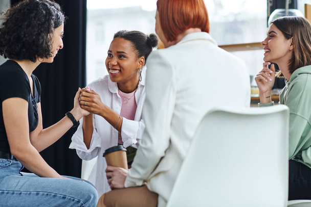 smiling african american woman holding hands with multiracial girlfriend near redhead motivation coach sitting with takeaway coffee on blurred foreground, support and mental health concept - Photo, Image