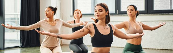 diverse multicultural female friends in sportswear practicing yoga and meditating with closed eyes and outstretched hands, inner peace and body awareness concept, banner - Zdjęcie, obraz