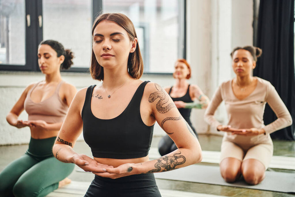young and tattooed woman in sportswear meditating with closed eyes near diverse group of multiethnic friends on blurred background, inner peace and body awareness concept - Photo, Image
