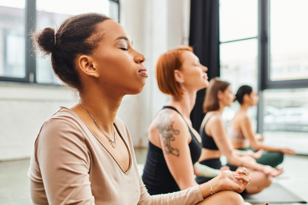 Genç Afrikalı Amerikalı kadın, yoga, ahenk ve refah konsepti sırasında rahat poz veren çok ırklı arkadaşlarının yanında, gözleri kapalı meditasyon yapıyor. - Fotoğraf, Görsel