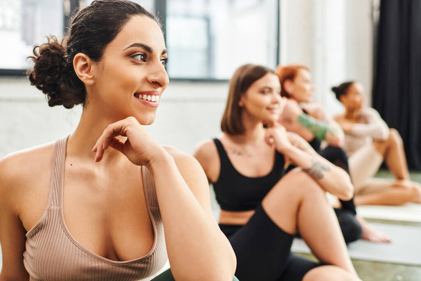 cheerful multiracial woman looking away during yoga class near diverse group of multiethnic girlfriends on blurred background, wellness and mental health concept - Photo, Image