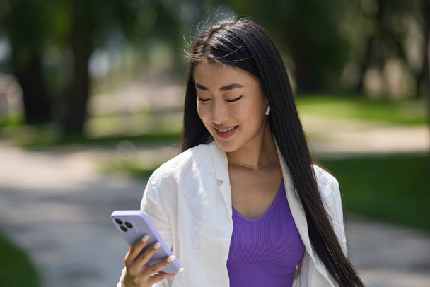 Portrait of a beautiful young Asian woman browsing songs catalog at online streaming app. Cheerful POC female using modern smart phone with a smile - Zdjęcie, obraz