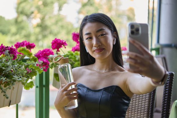 Cheerful Asian woman taking a selfie with a glass bottle of mineral water. Portrait of a beautiful POC female from Vietnam posing for a selfie photo in a cafe - Photo, image