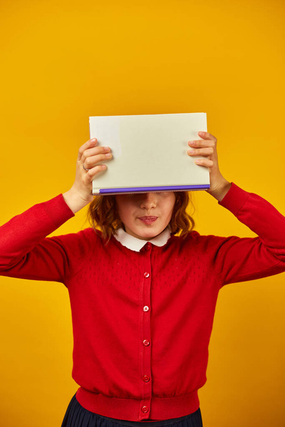 Portrait of happy teenage schoolgirl cover her face by book on yellow studio background, educatin concept, having fun , Back to School, copy space. - Foto, Imagem