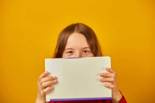 Portret van gelukkige tiener schoolmeisje cover haar gezicht door boek over gele studio achtergrond, educatin concept, plezier hebben, Terug naar School, kopieer ruimte. - Foto, afbeelding