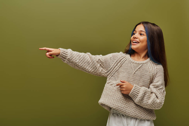 Веселая и модная девочка-подросток с крашеными волосами в современном трикотажном свитере и с пальцем, изолированным на зеленом, современном стиле для подростковой концепции - Фото, изображение
