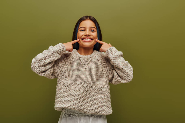 Smiling brunette preteen girl with dyed hair wearing stylish knitted sweater while pointing with finger at mouth and standing isolated on green, fashion-forward preteen with sense of style - Photo, Image
