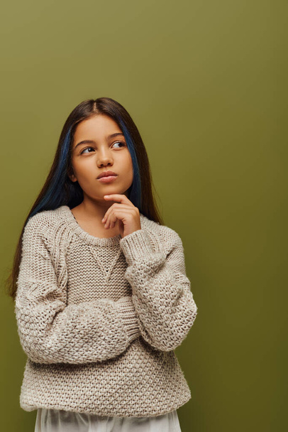 Pensive brunetka preteen dívka s barvenými vlasy na sobě stylový pletený svetr a dívá se stranou, zatímco stojí izolované na zelené, módní pre-teen se smyslem pro styl - Fotografie, Obrázek
