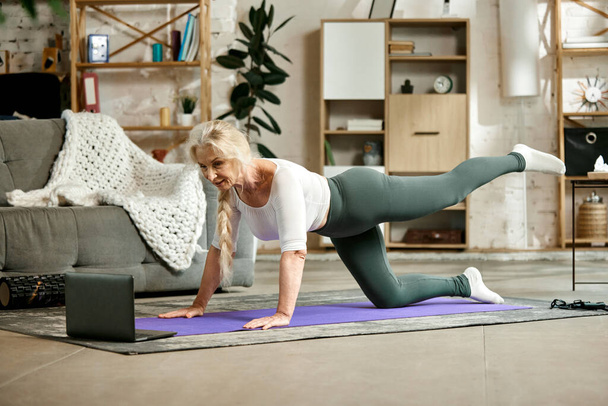 Beautiful, positive, senior woman in comfortable sportswear training at home with online fitness lessons via laptop. Concept of sportive and healthy lifestyle, age, body care, fitness, wellness. - 写真・画像