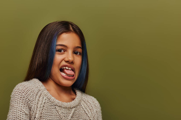 Портрет стильной девочки-подростка с окрашенными волосами в трикотажный свитер и торчащим языком при взгляде на камеру, изолированную на зеленом, современном и модном дизайне - Фото, изображение