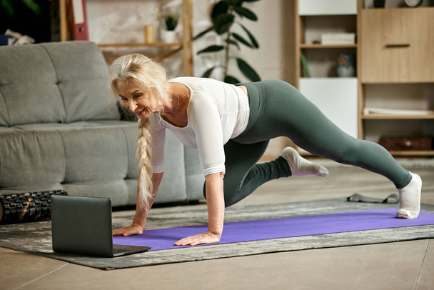 Beautiful, positive, senior woman in comfortable sportswear training at home with online fitness lessons via laptop. Concept of sportive and healthy lifestyle, age, body care, fitness, wellness. - Fotoğraf, Görsel