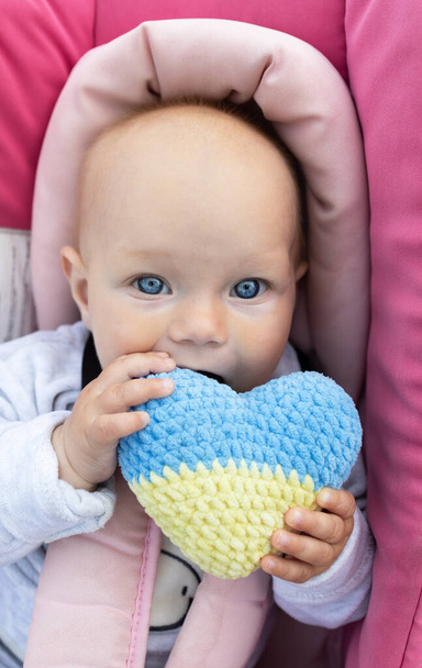 αθώο μωρό με μια εκφραστική εμφάνιση κρατά ένα πλεκτό κίτρινο-μπλε ουκρανική καρδιά στα χέρια. Ζητείται από τα παιδιά να σταματήσουν τον πόλεμο στην Ουκρανία. Πρόσφυγες με αγάπη για την Ουκρανία. Ημέρα ανεξαρτησίας - Φωτογραφία, εικόνα