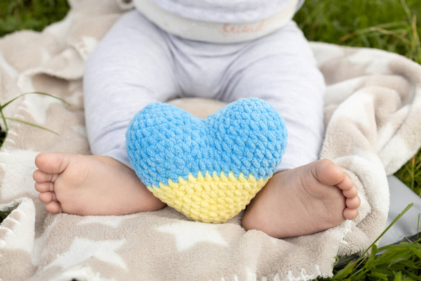 Yalın ayak bir bebek ve örülmüş sarı-mavi bir Ukrayna kalbi. Çocuklardan Ukrayna 'daki savaşı durdurmaları isteniyor. Ukrayna 'yı seven mülteciler. Bağımsızlık Günü. Vatansever kavram - Fotoğraf, Görsel