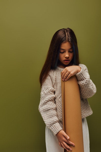 Стильный ребенок-подросток с окрашенными волосами, одетый в уютный и современный вязаный свитер, держа в руках бумагу, стоя изолированно на зеленом, девушка излучает осенние вибрации концепции - Фото, изображение