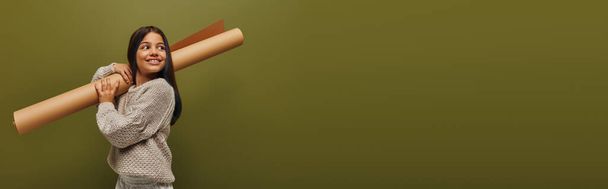 Glimlachend en trendy preadolescent in de herfst gebreide trui die wegkijkt terwijl hij opgerold papier vasthoudt en geïsoleerd staat op groen, meisje dat herfstvibes uitstraalt, banner met kopieerruimte - Foto, afbeelding