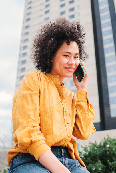 Retrato vertical de una joven latina con el pelo rizado teniendo una conversación telefónica con un smartphone. Señora hispana feliz con camisa amarilla hablando por teléfono celular sentado al aire libre. Mujer usando el teléfono - Foto, Imagen