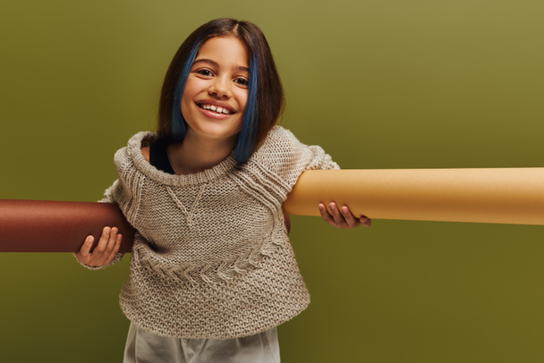 Positive preteen child with dyed hair wearing stylish knitted sweater and autumn outfit looking at camera and holding rolled papers isolated on green, autumn fashion for preteens concept - Photo, Image
