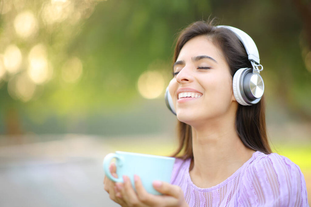 Happy woman wearing headphone listening to music holding coffee mug in a park and breathing fresh air - Foto, immagini