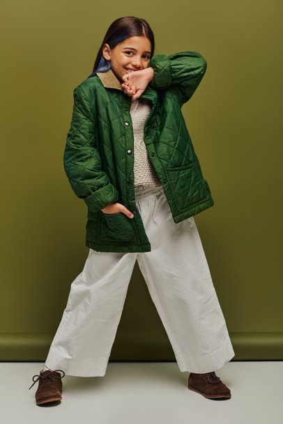 Volledige lengte van positieve pre-adolescent meisje met geverfd haar op zoek naar camera terwijl poseren in de herfst outfit en gebreide trui op groene achtergrond, moderne herfst mode voor pretiens concept - Foto, afbeelding
