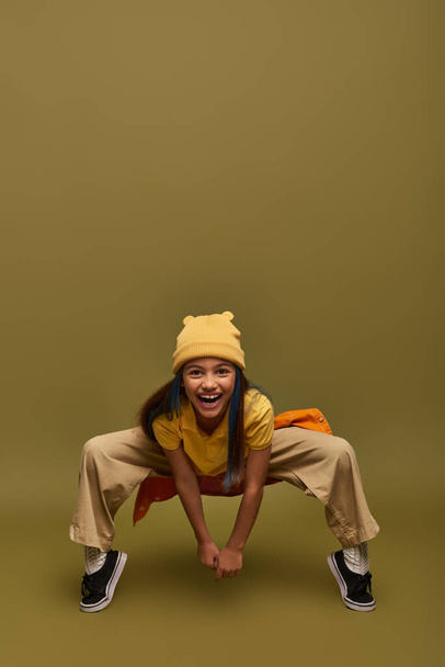 Full length of cheerful preadolescent girl with dyed hair wearing yellow hat and trendy clothes posing while looking at camera on khaki background, stylish girl in modern outfit concept - 写真・画像