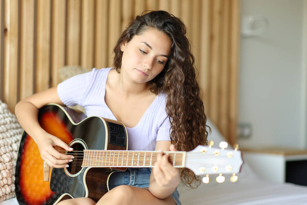 Woman playing guitar sitting on the bed in a room - Foto, afbeelding