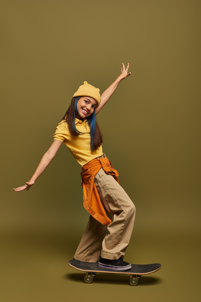 Positive and stylish preadolescent girl with dyed hair wearing yellow hat and urban outfit while looking at camera near skateboard on khaki background, stylish girl in modern outfit concept - Photo, Image