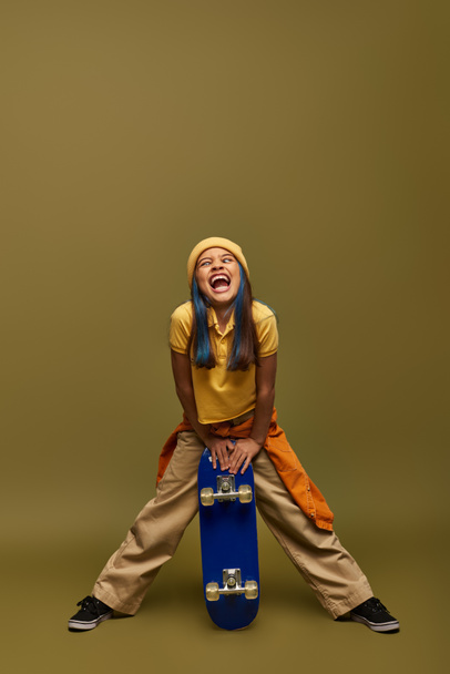 Volle Länge der aufgeregten Preteen Kind mit farbigen Haaren trägt urbane Outfit und Hut während Grimassen und hält Skateboard auf khakifarbenem Hintergrund, stilvolles Mädchen in modernem Outfit-Konzept - Foto, Bild