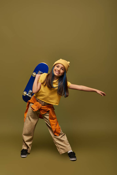 Full length of positive preteen girl with dyed hair wearing yellow hat and urban outfit while holding skateboard and standing on khaki background, stylish girl in modern outfit concept - Photo, Image