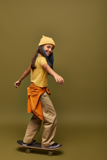 Smiling and trendy preadolescent girl with dyed hair in yellow hat and urban outfit looking at camera and standing on skateboard on khaki background, girl with cool street style look - Fotografie, Obrázek