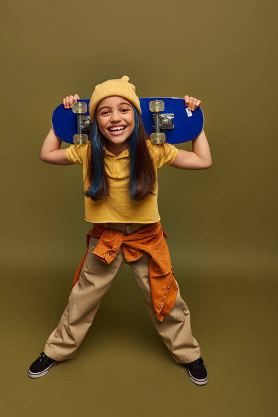 Volle Länge des lächelnden Frühchens mit gefärbten Haaren, gelbem Hut und urbanem Outfit, das Skateboard hält und auf khakifarbenem Hintergrund in die Kamera schaut, Mädchen mit coolem Street-Style-Look - Foto, Bild
