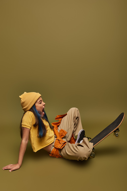 Side view of excited preadolescent girl with colorful hair wearing trendy urban outfit and hat while sitting near skateboard on khaki background, girl with cool street style look - Photo, Image
