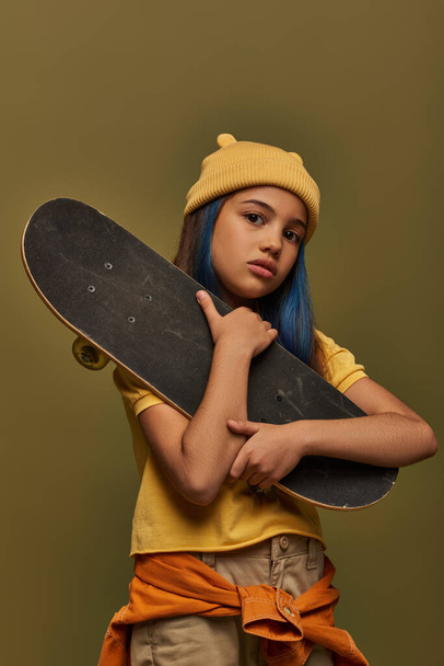 Portrait of stylish preadolescent girl in yellow hat and urban outfit looking at camera while holding skateboard and posing isolated on khaki, girl in urban streetwear concept - Foto, Imagem