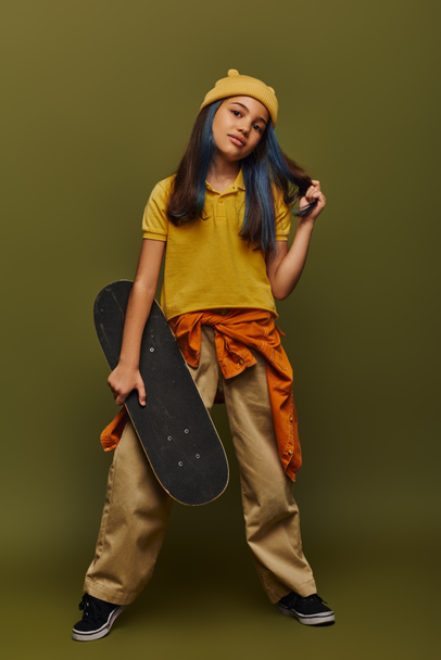 Volledige lengte van zelfverzekerde en modieuze tiener meisje met geverfd haar dragen hoed en stedelijke outfit en houden skateboard op kaki achtergrond, meisje in stedelijke streetwear concept - Foto, afbeelding