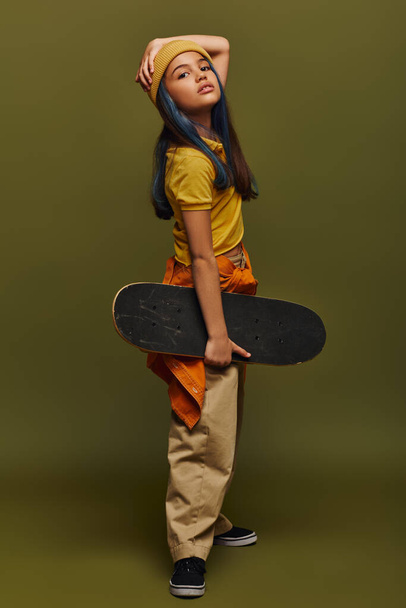 Volle Länge des trendigen und selbstbewussten vorpubertären Kindes mit farbigen Haaren, das Hut und urbanes Outfit trägt und Skateboard hält, während es auf khakifarbenem Hintergrund steht, Mädchen im urbanen Streetwear-Konzept - Foto, Bild